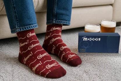 Ruby red Cheers beer socks with cream "Cheers!" script, worn with jeans on a grey rug beside two pints and the navy WearMoods gift box