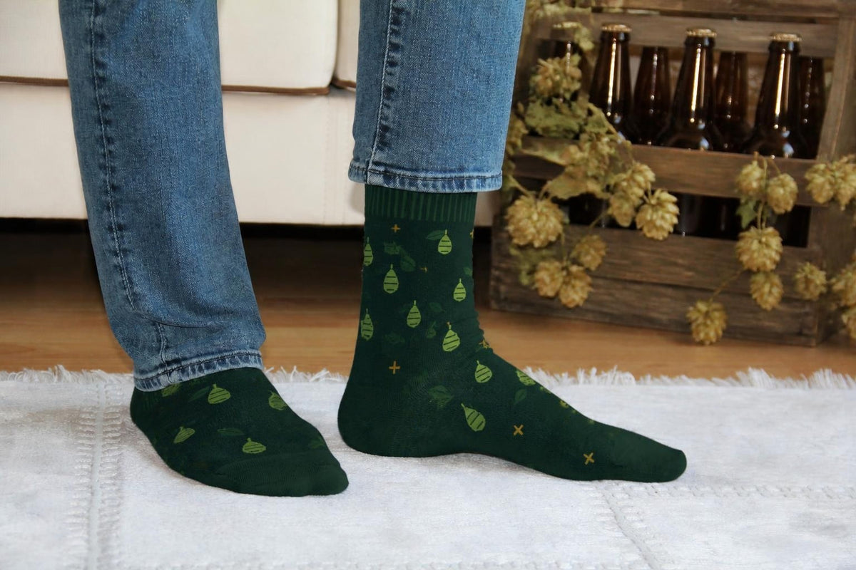 WearMoods Hop Field beer socks: forest green socks with hop cones, in front of a sofa and a crate of craft beer